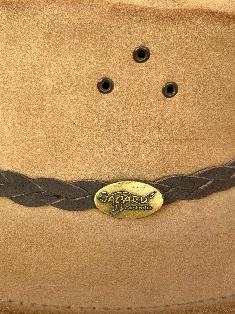 May include: A tan-colored Jacaru Australia hat with a braided band and metal emblem. The hat features three small eyelets on the crown. The hat is made of a suede-like material.