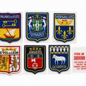 May include: Six embroidered souvenir patches featuring French city crests including Palavas les Flots, Dinard, Versailles, S'Pol de Leon, Angers, and Quimper, alongside a clear plastic instruction label with red text reading Stick on Souvenir Badge.