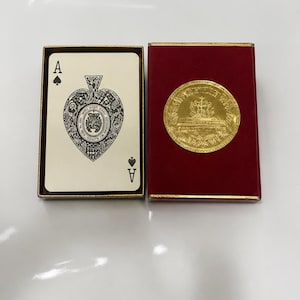 May include: A deck of playing cards in a gold-trimmed box with an ace of spades showing a detailed black and white crest. Beside it, a red velvet-textured box lid features a prominent circular gold medallion with an embossed crest and text.