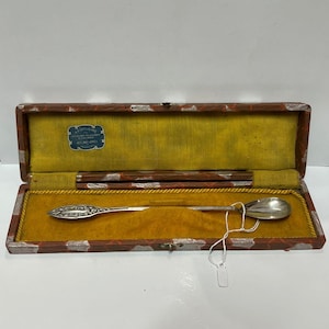 Pode incluir: Uma colher de prata com cabo e tigela ornamentados, apresentada numa caixa de madeira retangular. A caixa tem um exterior com padrão vermelho e branco, um interior amarelo e uma etiqueta. A colher tem cerca de 20 cm de comprimento.