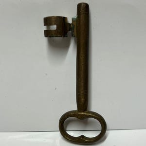 Può includere: Un'antica chiave in ottone con testa rettangolare e lungo stelo. La chiave ha un manico ovale con un ritaglio decorativo. L'ottone ha un aspetto ossidato e invecchiato.