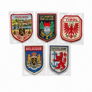 Könnte beinhalten: Fünf bestickte schildförmige Aufnäher in einzelnen Kunststoffhüllen. Die Aufnäher zeigen farbenfrohe Designs und die Schriftzüge HEIDELBERG, LA CHARTRE SUR-LE-LOIR, TIROL, BELGIQUE und DUSSELDORF mit verschiedenen heraldischen Symbolen und Landschaften.