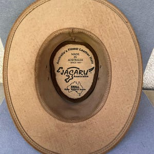 May include: Tan leather hat with a wide brim, viewed from the inside. The hat features a brown interior with a label that reads "Australia's Finest Leather Hats" and "JACARU Australia". The hat is labeled "SMALL" and "Style: Wallaroo Suede".