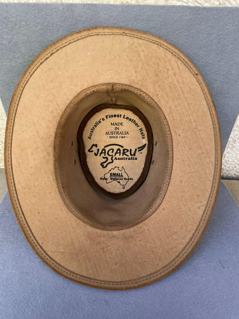 May include: Tan leather hat with a wide brim, viewed from the inside. The hat features a brown interior with a label that reads "Australia's Finest Leather Hats" and "JACARU Australia". The hat is labeled "SMALL" and "Style: Wallaroo Suede".