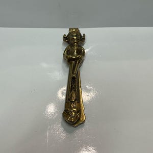 May include: A gold-toned metal door knocker shaped like a small person holding a staff. The knocker has a detailed design and a small, round face.