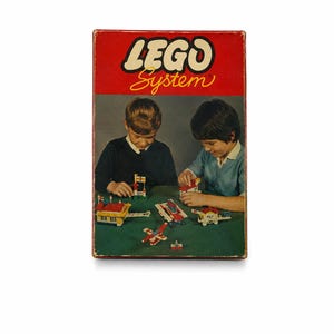 Pode incluir: Uma caixa de brinquedo vintage LEGO System com uma secção superior vermelha com o logótipo branco LEGO System. A parte inferior apresenta um gráfico de duas crianças a montar várias pequenas estruturas de blocos de plástico, incluindo uma casa e um avião.