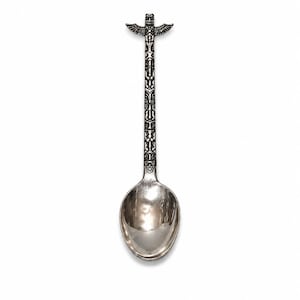 Op de afbeelding: Een zilverkleurige metalen lepel met een decoratieve steel voorzien van een ingewikkeld totempaalontwerp. De bovenkant van de steel is gevormd als een gevleugelde figuur, terwijl de steel herhalende snijpatronen vertoont. De lepelbak is glad.