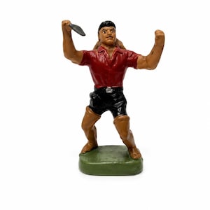 May include: A painted figurine of a person standing on a green base, wearing a red collared shirt and black shorts. The figure has dark hair, tan skin, and is holding a small gray object in the right hand while the left arm is raised in a flexed position.