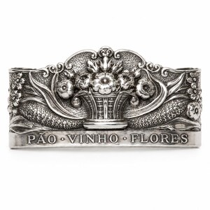 Puede incluir: Un servilletero de metal plateado con un diseño en relieve de una cesta de flores central rodeada de motivos florales y hojas texturizadas. La parte frontal del anillo muestra el texto grabado PÃO • VINHO • FLORES en letras mayúsculas.