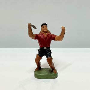 May include: A small, vintage figurine of a man in a red shirt, black shorts, and brown boots, holding a silver object. The figurine stands on a green base. The figure is in a throwing pose.