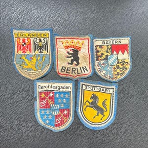 Könnte beinhalten: Fünf Vintage-Aufnäher mit Stadtwappen. Die Aufnäher zeigen Erlangen, Berlin, Bayern, Berchtesgaden und Stuttgart. Jeder Aufnäher hat einen blauen Rand und zeigt detaillierte Stadtsymbole und Text.