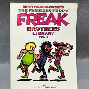 Puede incluir: Portada de cómic de "The Fabulous Furry Freak Brothers Library Vol. 1" de Gilbert Shelton. El título es rosa y rojo, con tres personajes de dibujos animados corriendo en la parte inferior. La portada es blanca.