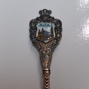 May include: A silver-toned souvenir spoon featuring a detailed shield design with the text "WIEN STEPHANSDOM" and an image of St. Stephen's Cathedral. The spoon has an ornate handle with a decorative pattern.