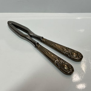 May include: A vintage silver-toned nutcracker with ornate handles. The nutcracker features a scissor-like design with serrated jaws for cracking nuts. The handles have a detailed, embossed pattern.
