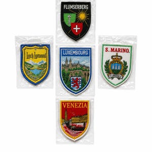 May include: Five shield-shaped embroidered patches in clear plastic sleeves. The patches feature designs for Flumserberg, Loch Lomond, Luxembourg, S. Marino, and Venezia, each displaying colorful regional imagery, text, and heraldic symbols.