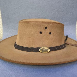 May include: A tan-colored hat with a wide brim and a braided dark brown band. The hat has three small air vents and a metal oval emblem. The hat is likely made of suede or a similar material.