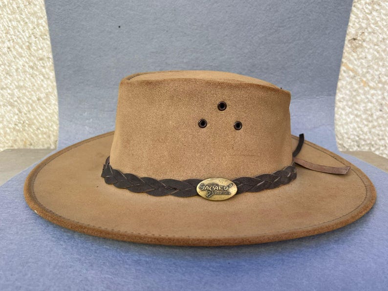 May include: A tan-colored hat with a wide brim and a braided dark brown band. The hat has three small air vents and a metal oval emblem. The hat is likely made of suede or a similar material.