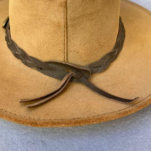 May include: A tan-colored hat with a wide brim and a braided leather band. The hat has a classic design, with a slightly curved brim and a structured crown. The leather band is dark brown and tied in a knot at the front.