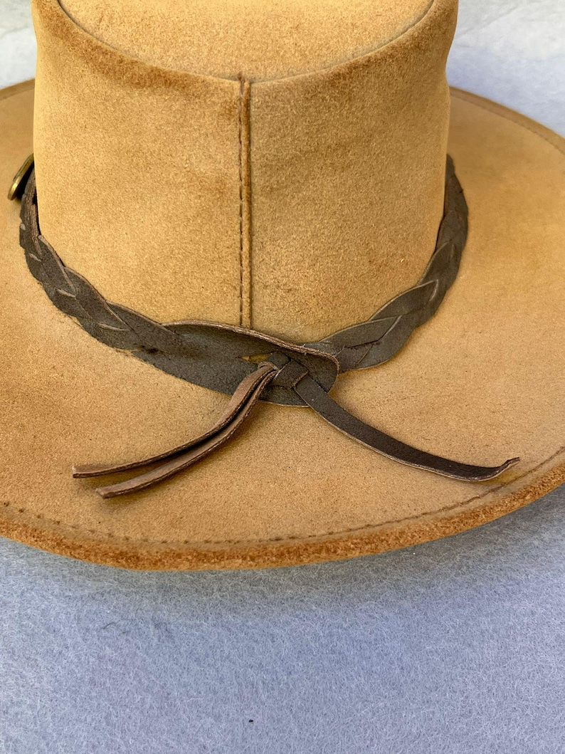May include: A tan-colored hat with a wide brim and a braided leather band. The hat has a classic design, with a slightly curved brim and a structured crown. The leather band is dark brown and tied in a knot at the front.