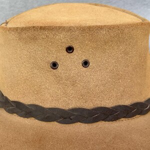 May include: A tan-colored hat with a braided dark brown band around the crown. The hat has three small, evenly spaced eyelets above the band. The hat's brim is slightly curved, and the material appears to be suede or a similar fabric.