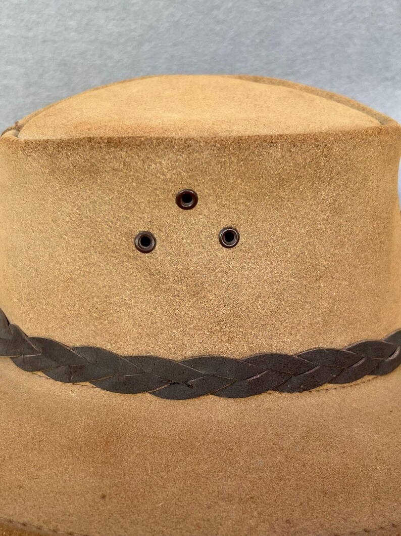May include: A tan-colored hat with a braided dark brown band around the crown. The hat has three small, evenly spaced eyelets above the band. The hat's brim is slightly curved, and the material appears to be suede or a similar fabric.