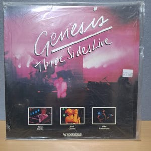 May include: A vinyl record album cover for Genesis's "Three Sides Live" album. The cover features a pink background with the band's name and album title in white text. The album cover also features photos of the band members: Tony Banks, Phil Collins, and Mike Rutherford.