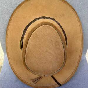 May include: A tan-colored cowboy hat with a rounded crown and wide brim. The hat features a dark brown braided band around the crown and a small decorative button. A leather tie hangs from the side of the hat.