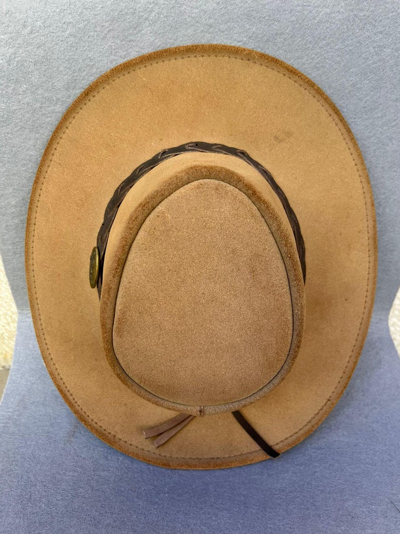 May include: A tan-colored cowboy hat with a rounded crown and wide brim. The hat features a dark brown braided band around the crown and a small decorative button. A leather tie hangs from the side of the hat.
