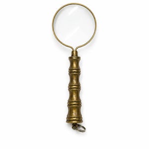 Pode incluir: Uma lupa com uma lente circular emoldurada em latão. O cabo é feito de latão com uma textura segmentada semelhante ao bambu. Um pequeno anel de metal está preso à base do cabo para permitir que o objeto seja pendurado ou fixado.