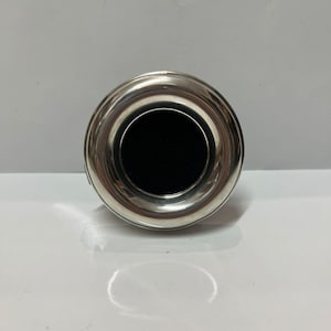 Pode incluir: Uma rolha de garrafa prateada com um design circular. A rolha tem um acabamento metálico brilhante e um inserto preto circular no centro. O design é simples e elegante.