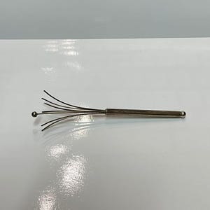 May include: A silver-coloured head massager with flexible wires and a rounded handle. The massager has multiple wires that extend outwards, ending in small, rounded tips. The handle is cylindrical and smooth.