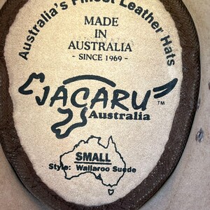 May include: Close-up of a leather hat label. The label is tan with dark brown text that reads "Australia's Finest Leather Hats, Made in Australia - Since 1969." The brand name "Jacaru Australia" is also visible, along with "Small" and "Wallaroo Suede."