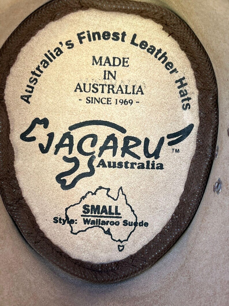 May include: Close-up of a leather hat label. The label is tan with dark brown text that reads "Australia's Finest Leather Hats, Made in Australia - Since 1969." The brand name "Jacaru Australia" is also visible, along with "Small" and "Wallaroo Suede."