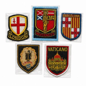 Puede incluir: Cinco parches bordados con escudos y monumentos de ciudades. Los parches muestran los nombres London, Inverness, Barcelona, München y Vaticano, cada uno con diseños heráldicos, colores e ilustraciones arquitectónicas distintos en fundas de plástico individuales.