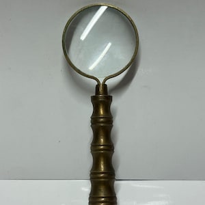 Pode incluir: Uma lupa de estilo vintage com uma lente clara e circular e uma alça de cor latão. A alça tem um design semelhante ao bambu e um pequeno anel é preso na base. A lupa foi projetada para leitura ou inspeção detalhada.
