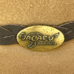May include: Close-up of a tan-colored hat with a dark brown leather band. The hat features a gold-colored oval emblem with the word "JACARU" and "Australia" inscribed on it.