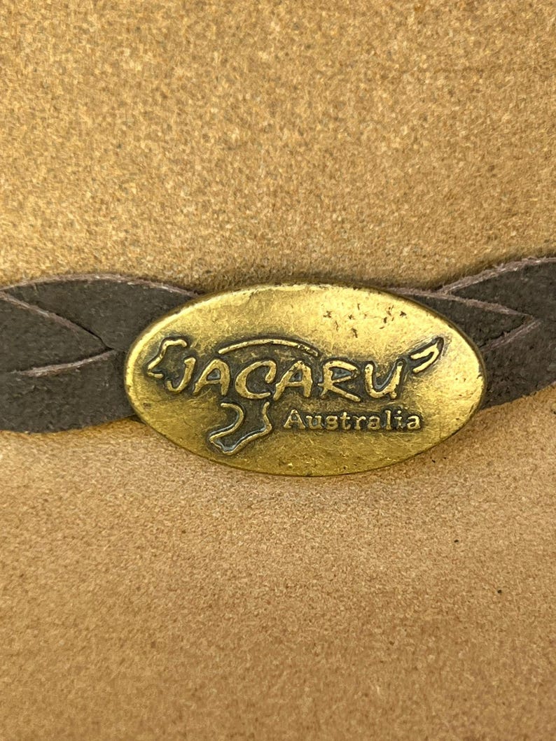 May include: Close-up of a tan-colored hat with a dark brown leather band. The hat features a gold-colored oval emblem with the word "JACARU" and "Australia" inscribed on it.