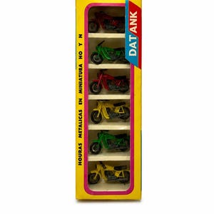 Puede incluir: Una caja de cartón amarilla vertical que contiene seis motocicletas de juguete en miniatura de colores rojo, verde y amarillo. El empaque presenta el texto "mini Moto" arriba, "DAT ANK" en el lateral y "HOY N HOURAS METALICAS EN MINIATURA" impreso verticalmente.