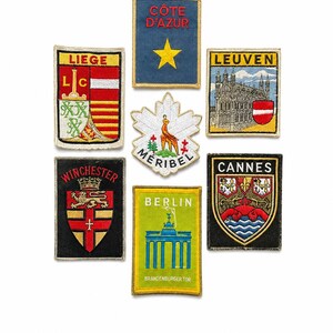 Op de afbeelding: Een verzameling van zeven geborduurde souvenirpatches met diverse locaties waaronder Côte d'Azur, Luik, Leuven, Méribel, Winchester, Berlijn en Cannes. Elke rechthoekige of gevormde patch toont kleurrijke regionale emblemen en tekst.
