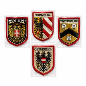 May include: Four embroidered shield-shaped patches with red borders. The patches feature the text WIEN, NURNBERG, STRATFORD UPON AVON, and ÖSTERREICH, along with heraldic designs including eagles, a chevron with bear heads, and diagonal stripes.
