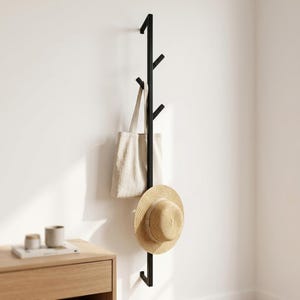 May include: A vertical black metal coat rack with multiple angled hooks mounted on a wall. A beige fabric tote bag hangs from one of the upper hooks, and a tan straw hat with a wide brim is suspended from a lower hook on the rack.