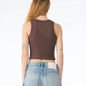 May include: A brown ribbed tank top with a scoop neckline. The cropped top is paired with light blue denim jeans. The model is facing away from the camera.