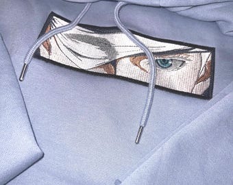 May include: A light blue hooded sweatshirt featuring a rectangular embroidered patch on the chest. The patch displays a detailed graphic of a single blue eye with white hair and a black border. The hoodie includes matching light blue drawstrings.