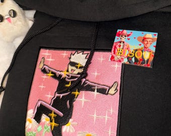 May include: A black hooded sweatshirt featuring a large square embroidered patch on the front. The patch depicts an anime character with white hair and a blindfold, surrounded by yellow sparkles and white daisies on a pink background.