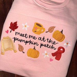 Puede incluir: Una sudadera rosa claro con bordados de temática otoñal que incluyen calabazas, un suéter, una taza, hojas, flores y una bufanda roja. El texto "meet me at the pumpkin patch" está bordado en cursiva negra en el centro del diseño.