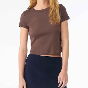 May include: A short-sleeved, brown crop top with a crew neck. The top is made of a solid, opaque material. The model is wearing a navy blue skirt.