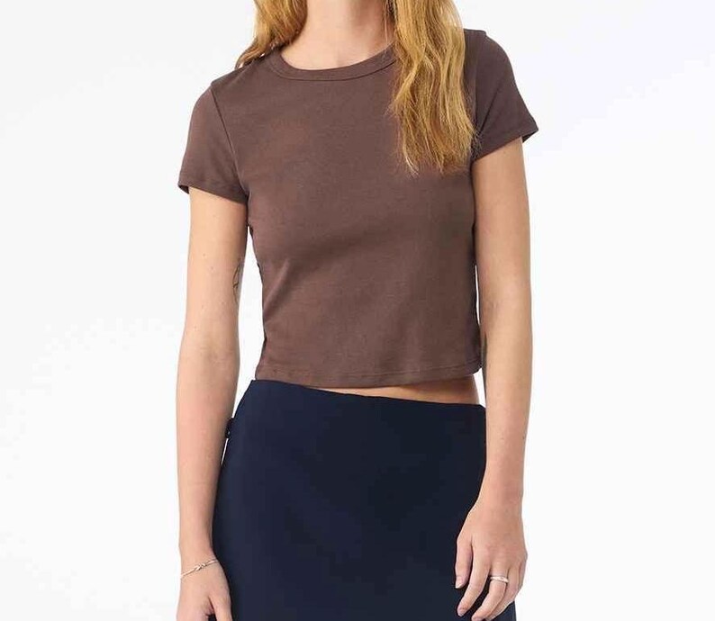 May include: A short-sleeved, brown crop top with a crew neck. The top is made of a solid, opaque material. The model is wearing a navy blue skirt.