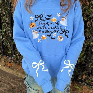 Könnte beinhalten: Ein hellblaues Sweatshirt mit einem gestickten Design und dem Text "big fan of little treats & halloween". Die Grafik zeigt Geister, Kürbisse, Tortenstücke, Fledermäuse, Blumen und schwarze Schleifen. Auf beiden Ärmeln sind weiße Schleifen aufgedruckt.