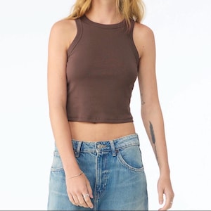 May include: A brown, cropped tank top with a ribbed texture. The top has a round neckline and a sleeveless design. The model is wearing blue jeans. The top is a solid color and appears to be made of a soft material.