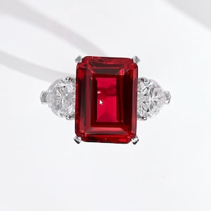 Puede incluir: Un lujoso anillo con una gran gema rectangular de color rojo intenso como pieza central, flanqueada por dos gemas claras en forma de pera. El anillo está engarzado en metal plateado o blanco, mostrando un diseño clásico y elegante.